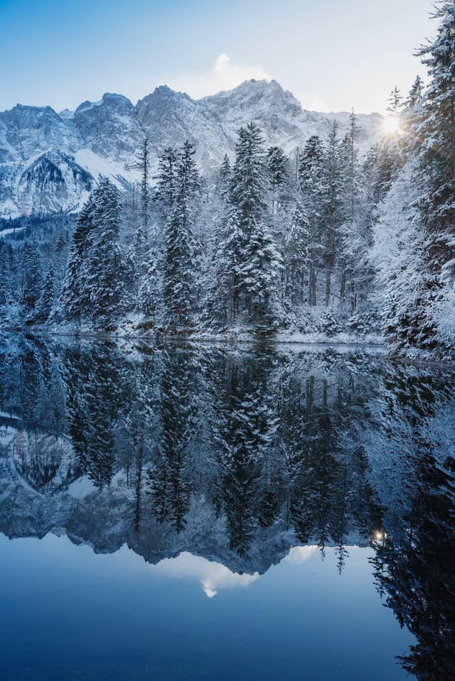 A serene winter landscape with snow-covered trees reflected in a calm lake, set against a backdrop of majestic mountains and a bright sky