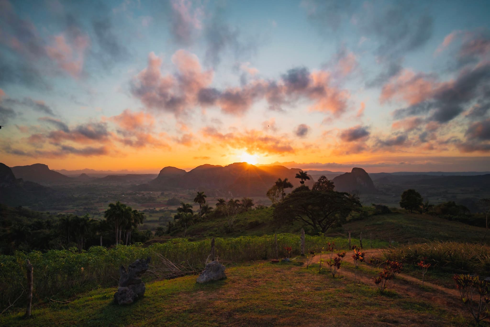 Colorful sunrise over a lush green valley with scattered trees, distant mountains, and a dramatic cloud-filled sky