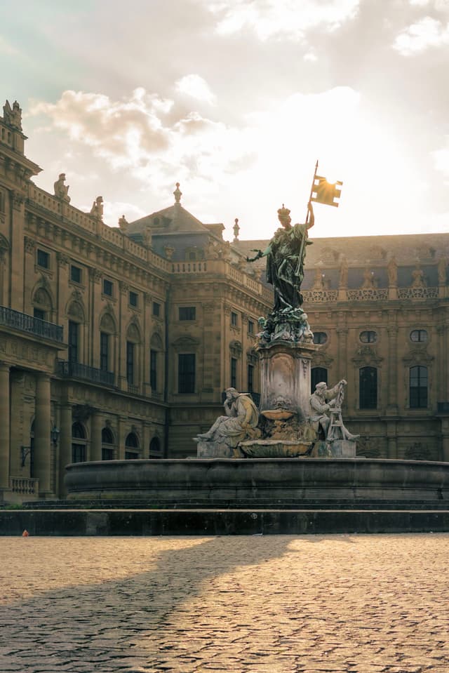 A grand statue stands in the center of a historic courtyard, surrounded by ornate architecture, with sunlight streaming through the clouds above