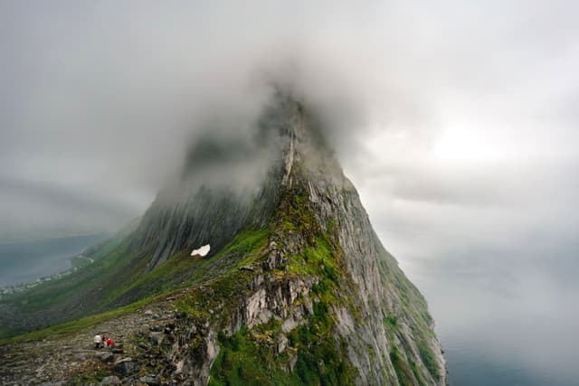 A steep, rocky mountain peak shrouded in mist, with patches of green vegetation and a cloudy sky