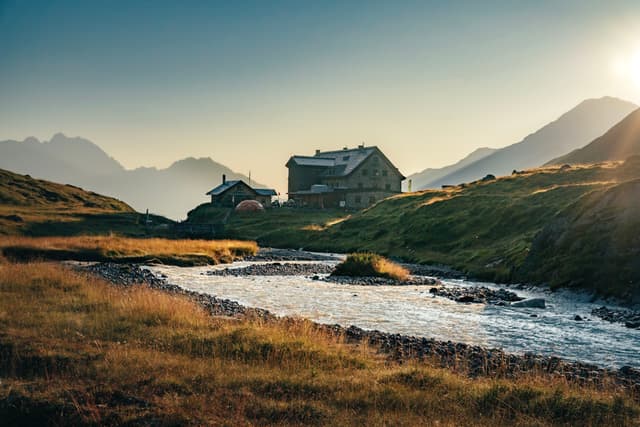 A serene mountain landscape with a river flowing through grassy fields, and rustic buildings nestled in the background under a clear sky