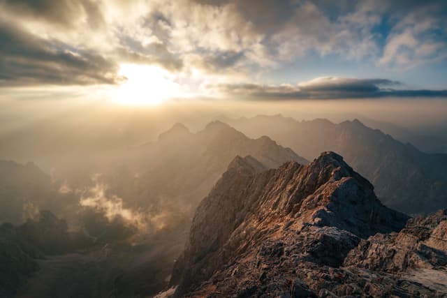 Sunset light streaming through clouds over a rugged mountain ridge with distant hazy peaks