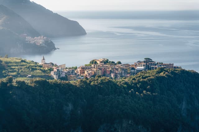 Hilltop coastal village with clustered pastel buildings and a church tower, surrounded by green terraces above a calm, hazy blue sea and distant mountains