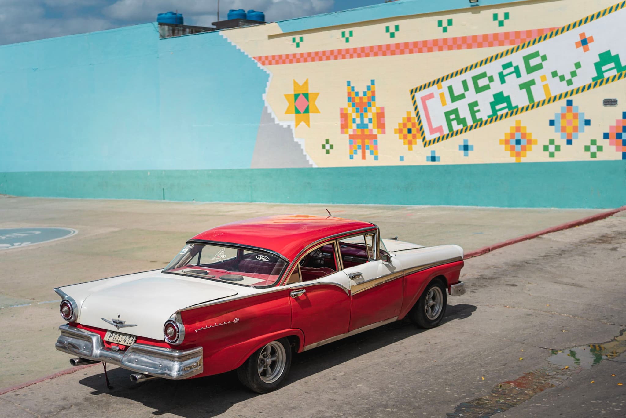 Red and white vintage car parked beside a colorful geometric mural on a sunny day