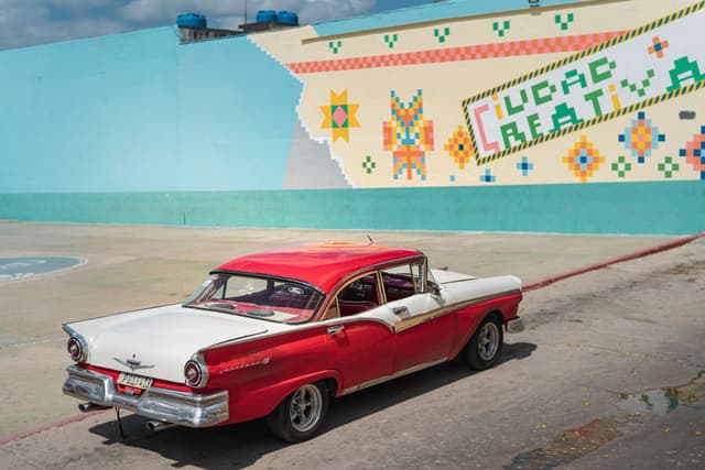 Red and white vintage car parked beside a colorful geometric mural on a sunny day