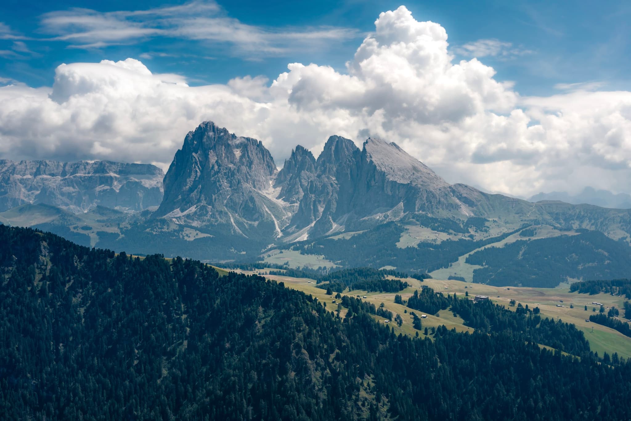 A majestic mountain range under a partly cloudy sky, with lush green valleys and forests in the foreground