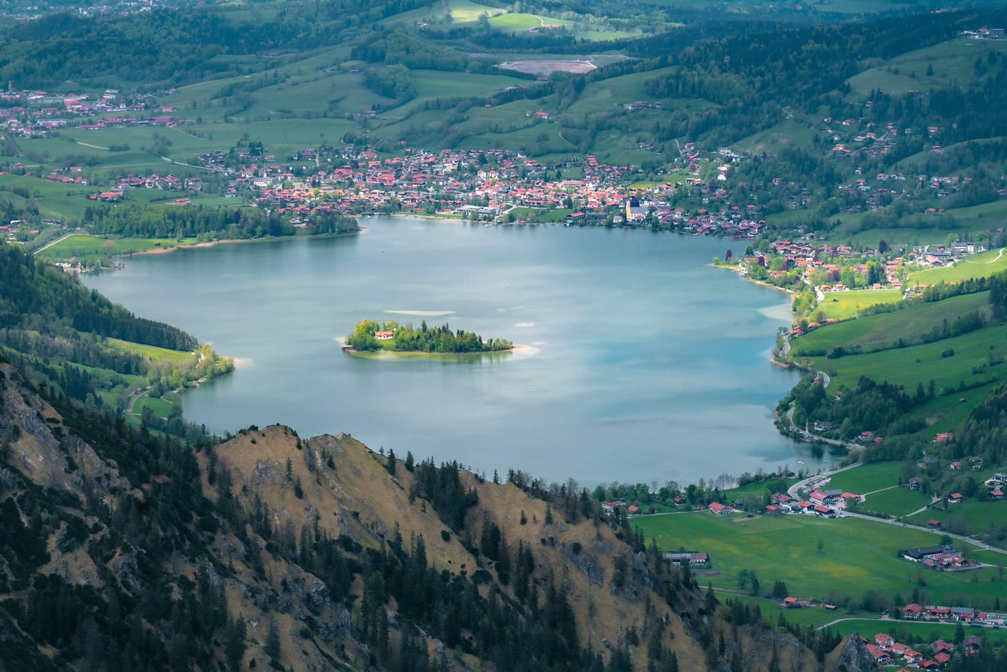 High-angle view of a mountain lake with a small central island, surrounded by green hillsides and a distant town along the shoreline