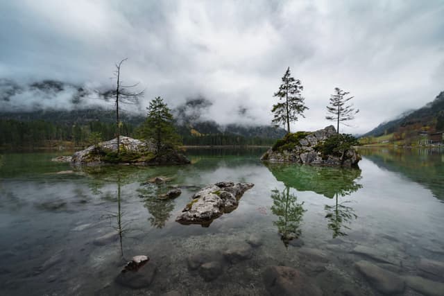 A serene lake with clear water reflecting trees and rocks, surrounded by misty mountains under a cloudy sky