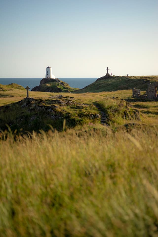 White lighthouse and stone cross standing on grassy coastal hills overlooking the sea