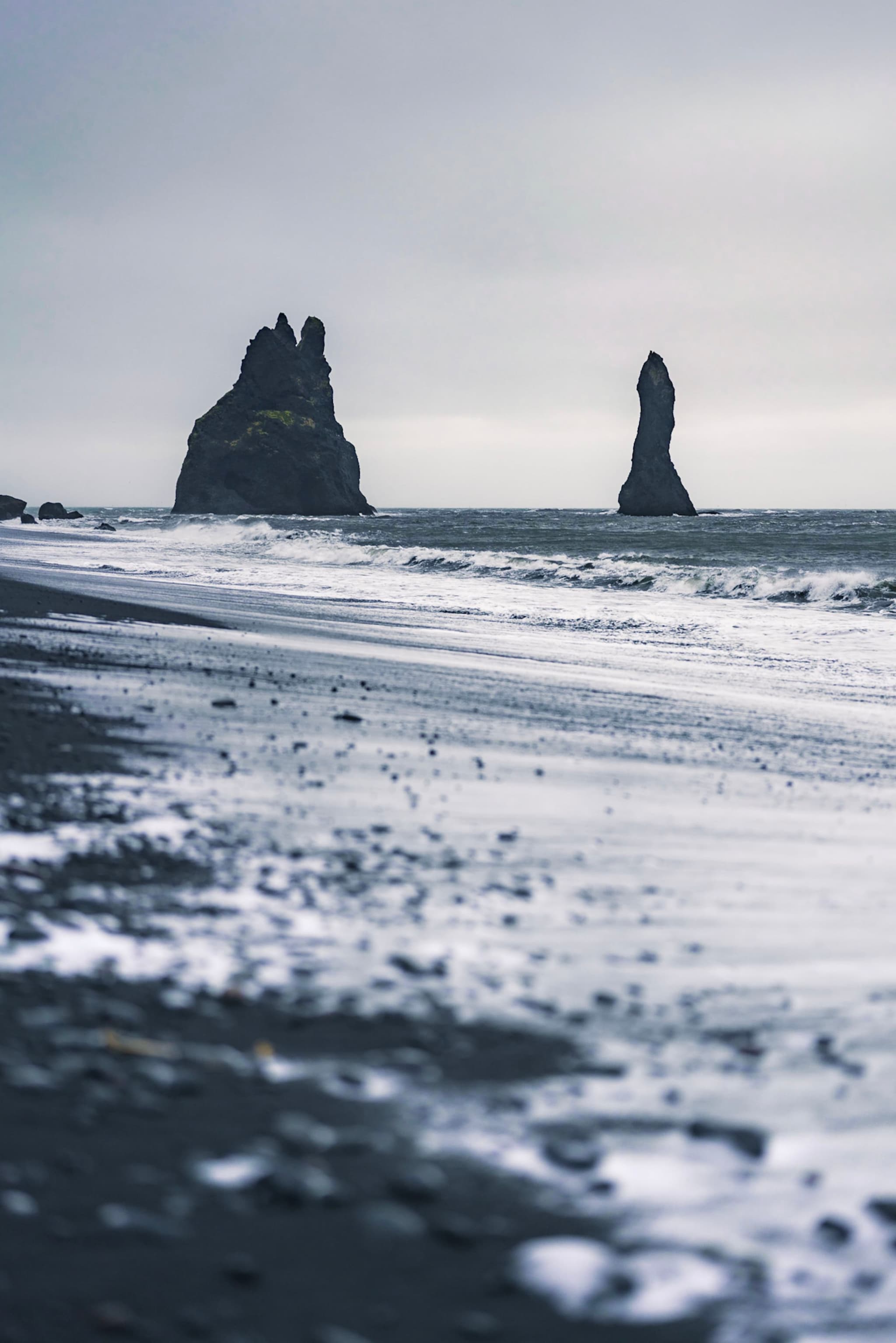 Moody seascape with dark sand beach, foamy waves, and tall rock stacks rising from the ocean under an overcast sky