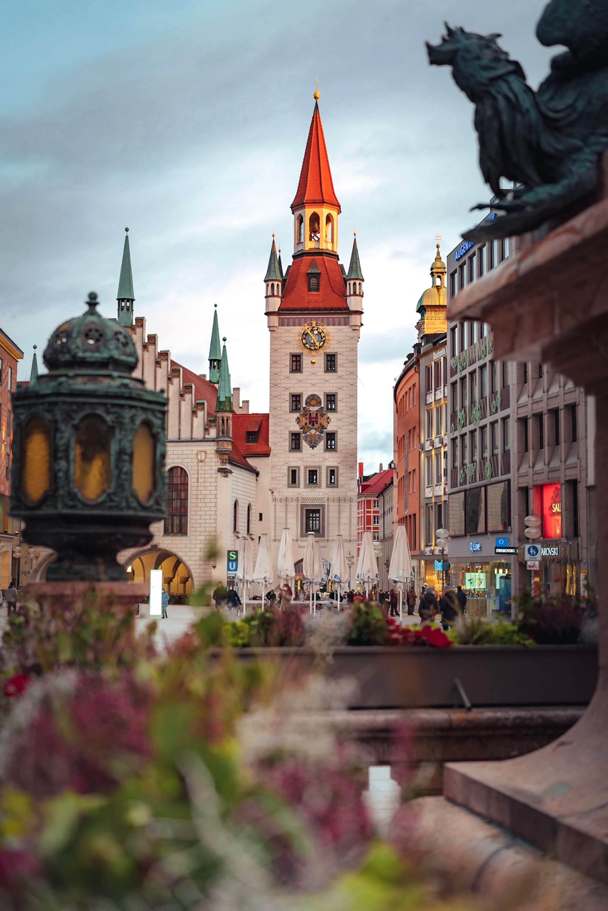 historic European street with central clock tower, surrounding buildings, and foreground flowers at dusk