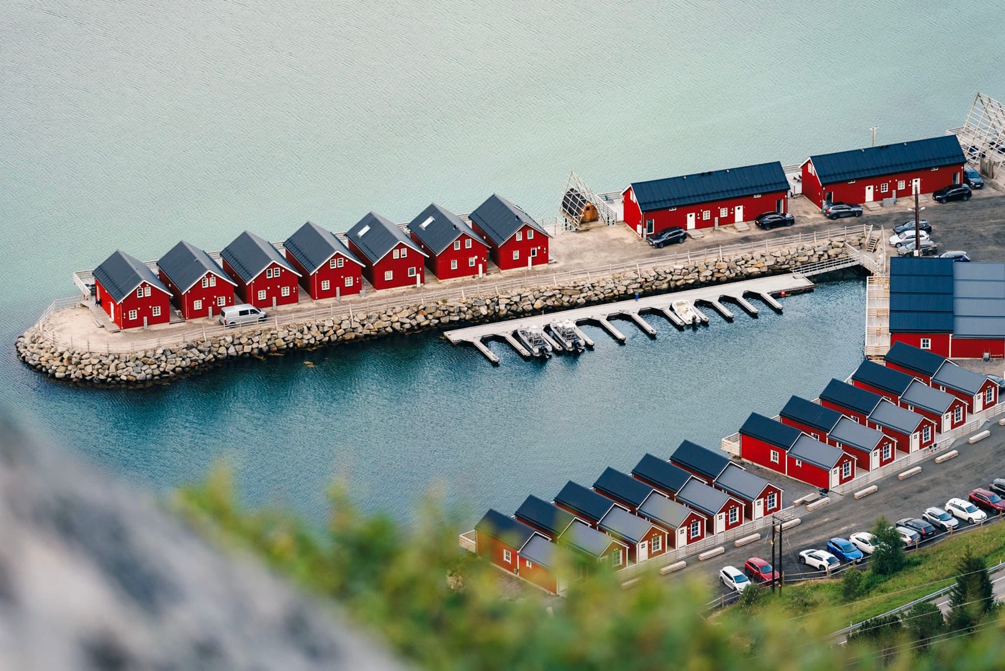 A row of red houses with black roofs lines a narrow strip of land extending into a body of water, with a small marina and boats docked nearby