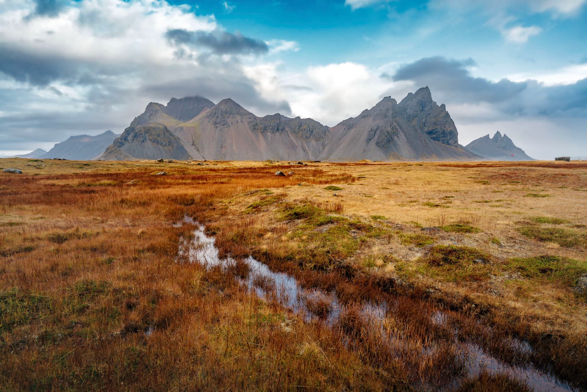 A landscape with rugged mountains in the background, a grassy field with patches of red and brown in the foreground, and a small stream running through the scene under a partly cloudy sky