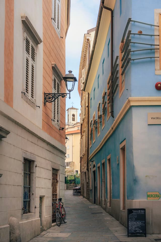 Narrow European alleyway between pastel buildings with shuttered windows, bicycles, and a distant church tower