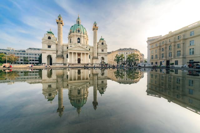 Baroque domed church and surrounding city buildings reflected symmetrically in a calm foreground pool under a bright sky