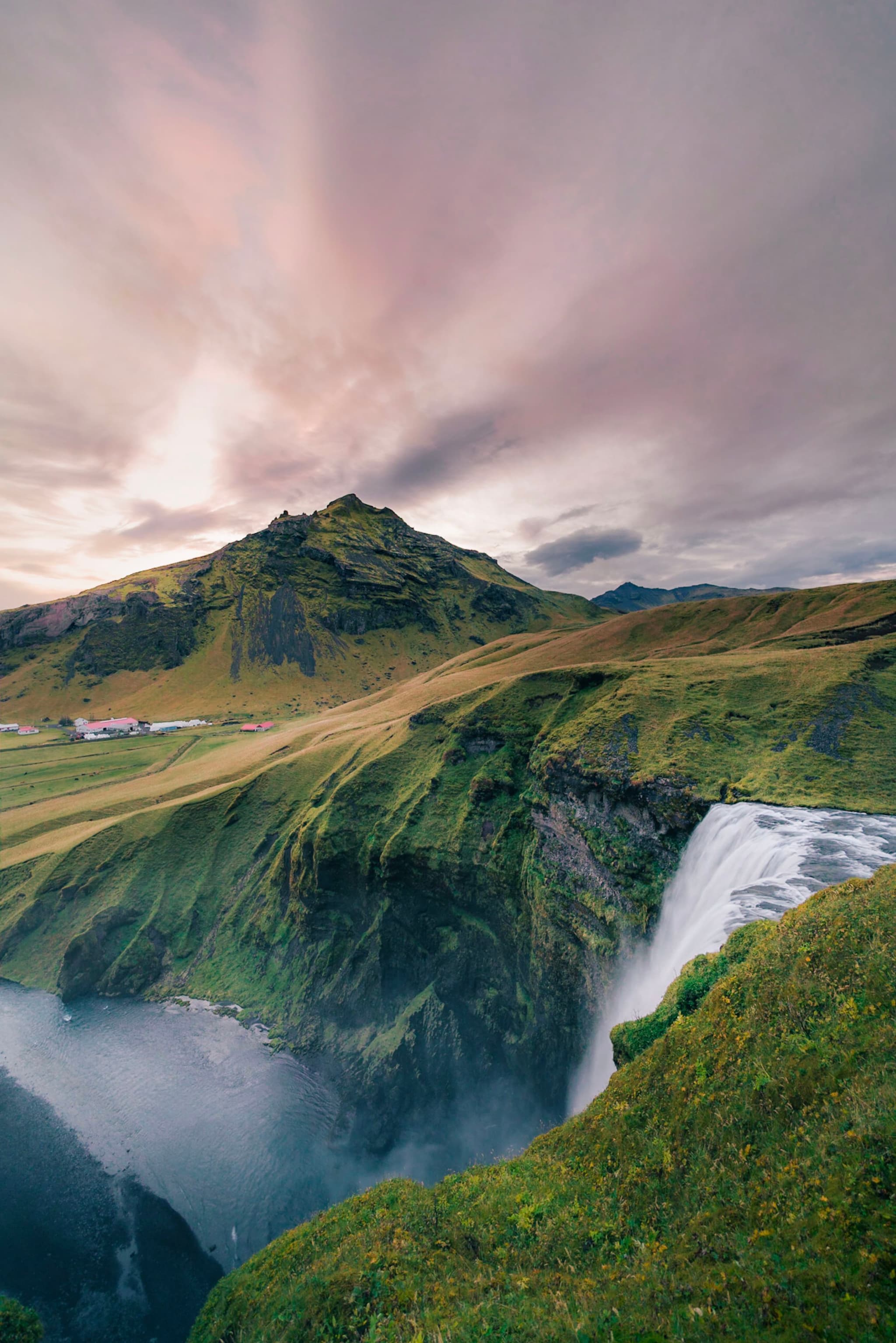 Tall waterfall plunging into a misty river gorge beneath a grassy, rugged mountain at dusk with streaked pastel clouds overhead