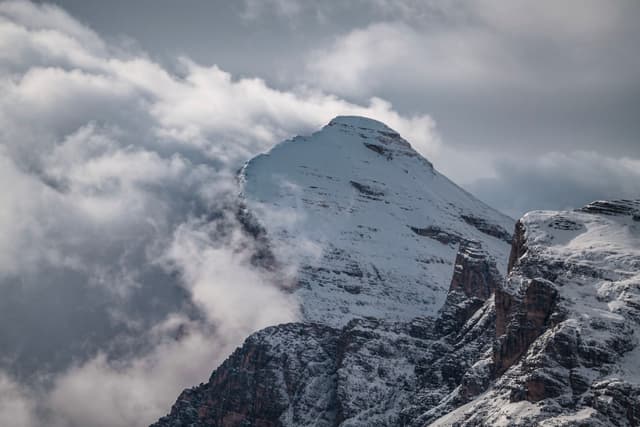 Snow-covered mountain peak partially obscured by swirling clouds above rugged rocky slopes
