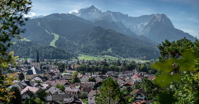 A picturesque town nestled in a valley with a backdrop of majestic mountains under a clear blue sky, surrounded by lush greenery