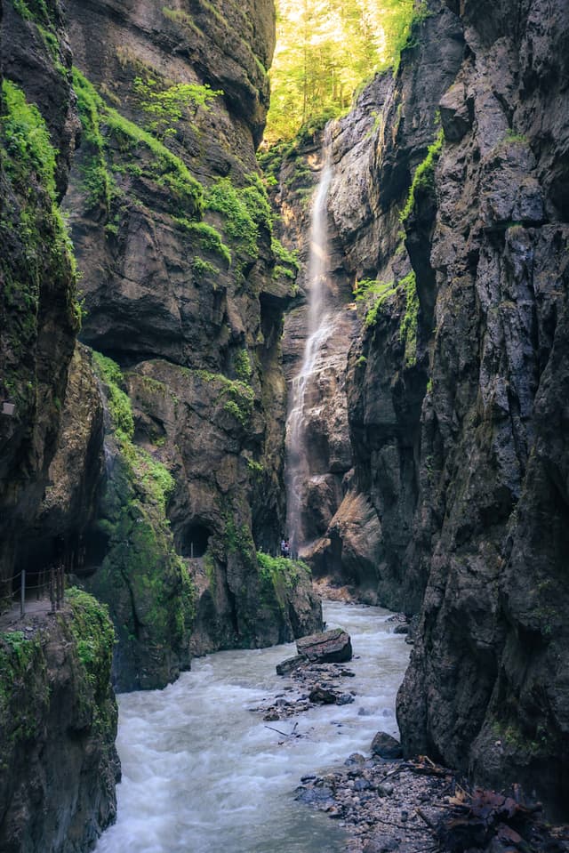 Narrow rock canyon with a tall waterfall cascading into a rushing stream between steep, mossy cliffs