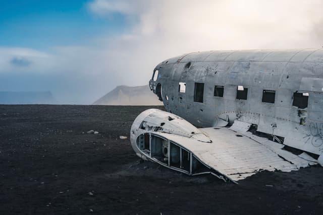 A weathered airplane wreck rests on a desolate, black sand landscape under a cloudy sky