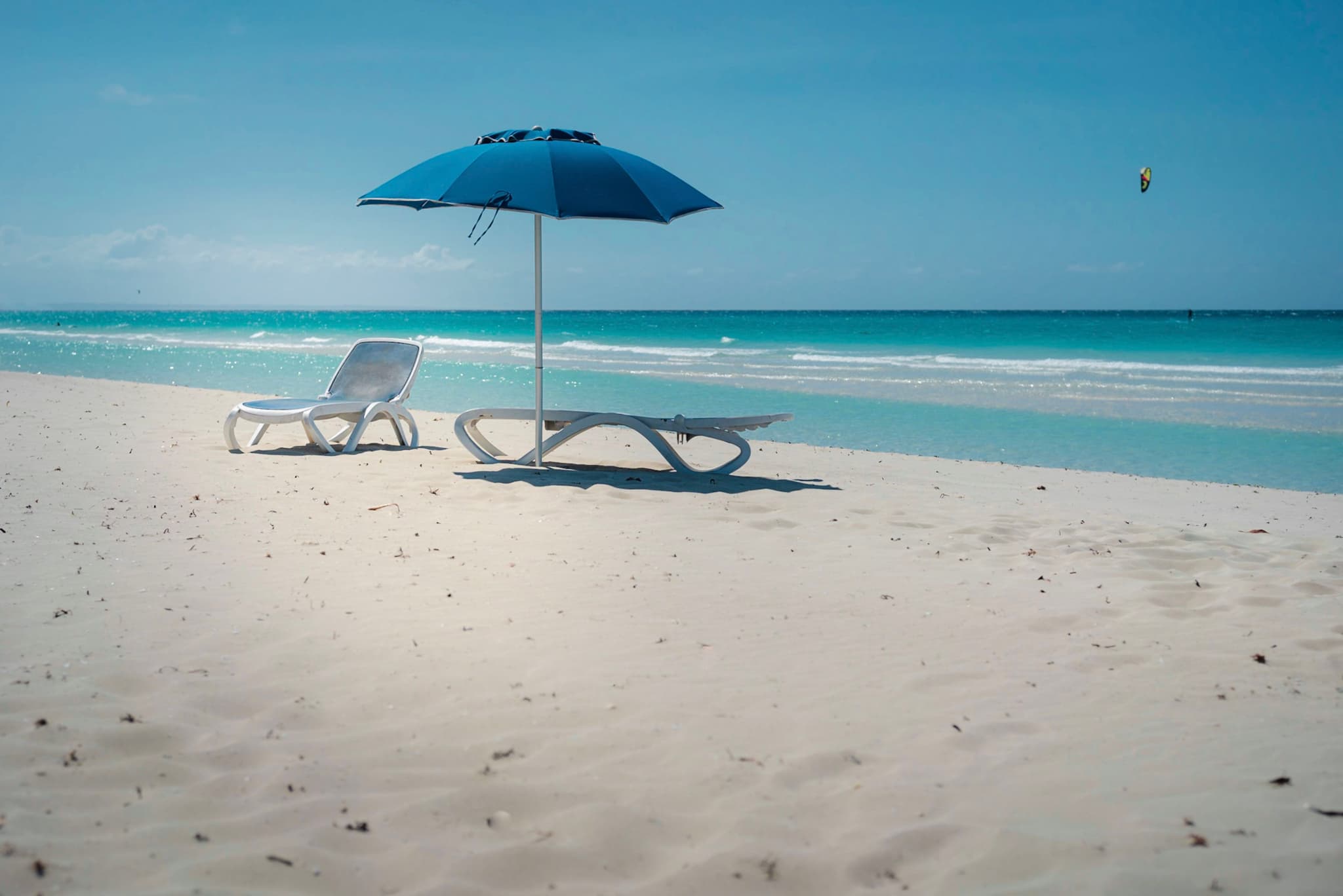 Two empty lounge chairs and a blue beach umbrella on a pristine sandy shore by a calm turquoise sea under a clear blue sky