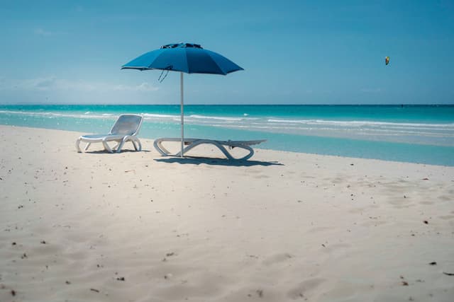 Two empty lounge chairs and a blue beach umbrella on a pristine sandy shore by a calm turquoise sea under a clear blue sky