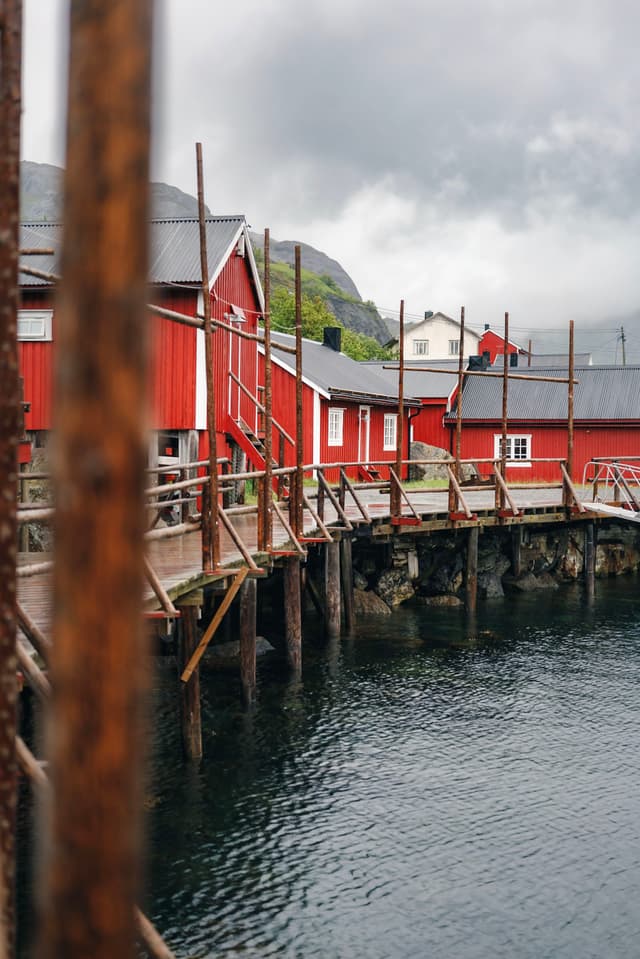 Red wooden buildings on stilts line a waterfront, with a cloudy sky and a mountainous backdrop