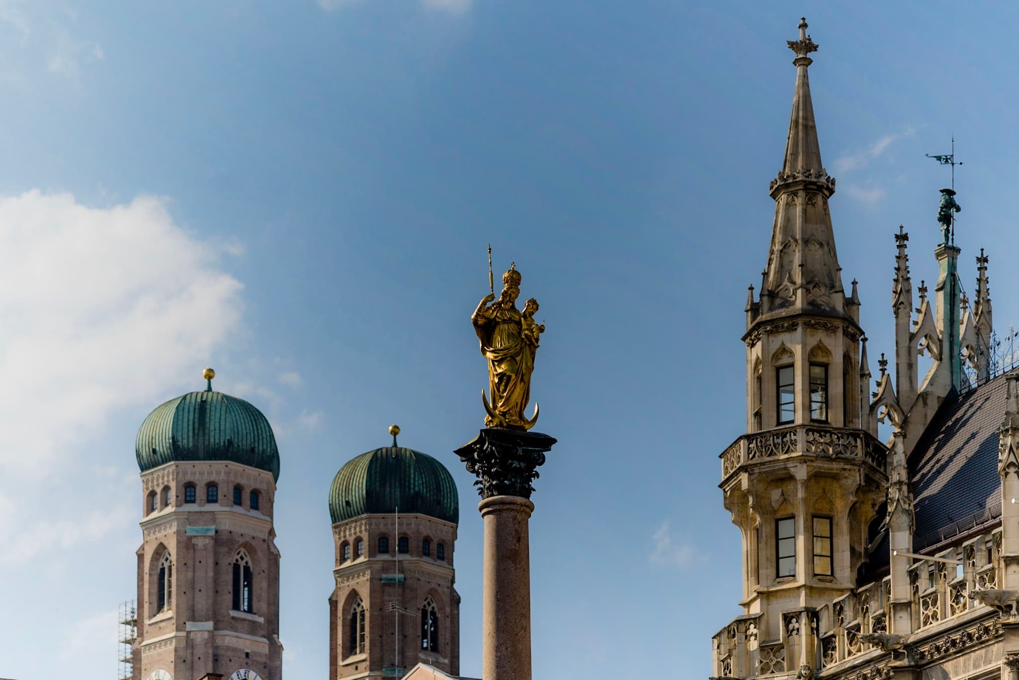 Golden statue atop a column framed by green-domed towers and ornate Gothic spire against a blue sky