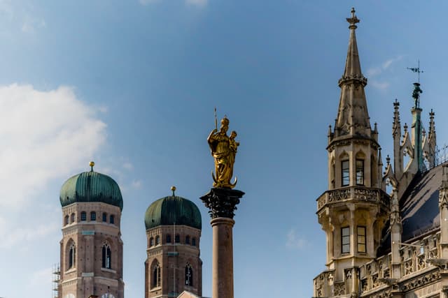 Golden statue atop a column framed by green-domed towers and ornate Gothic spire against a blue sky