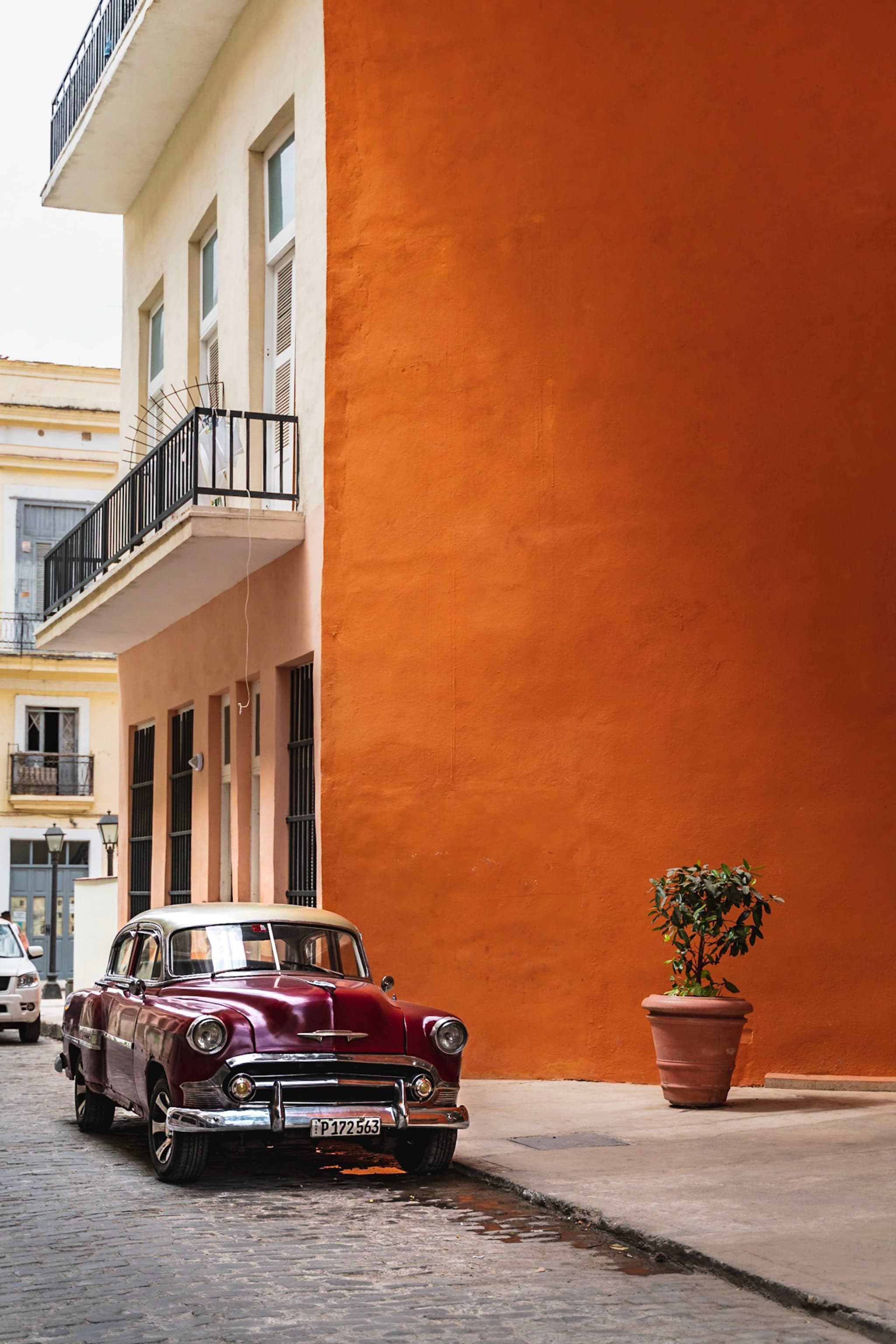Classic maroon car parked beside a tall orange wall on a narrow cobblestone street with a single potted plant