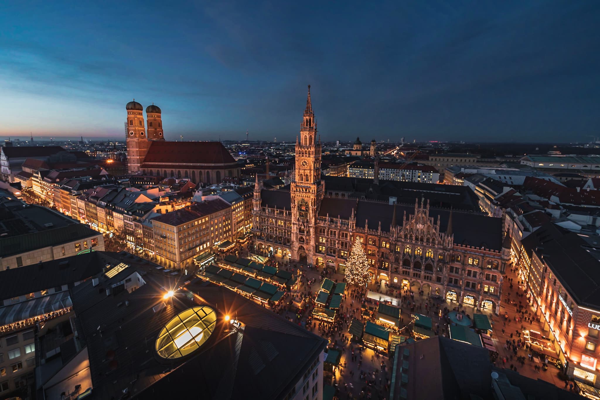 Elevated dusk view of a historic European city square with illuminated market stalls, a tall clock tower, and surrounding buildings glowing under a deep blue sky