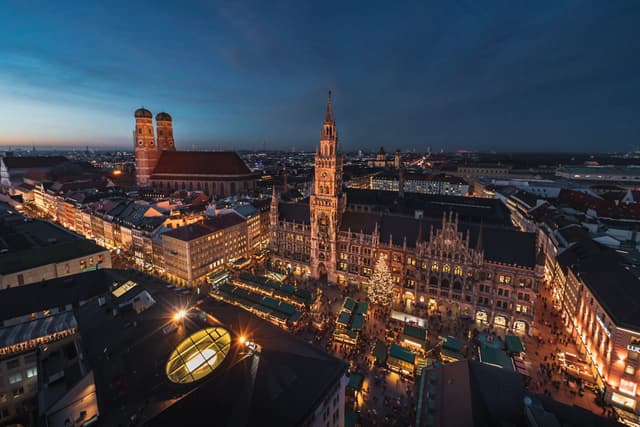 Elevated dusk view of a historic European city square with illuminated market stalls, a tall clock tower, and surrounding buildings glowing under a deep blue sky