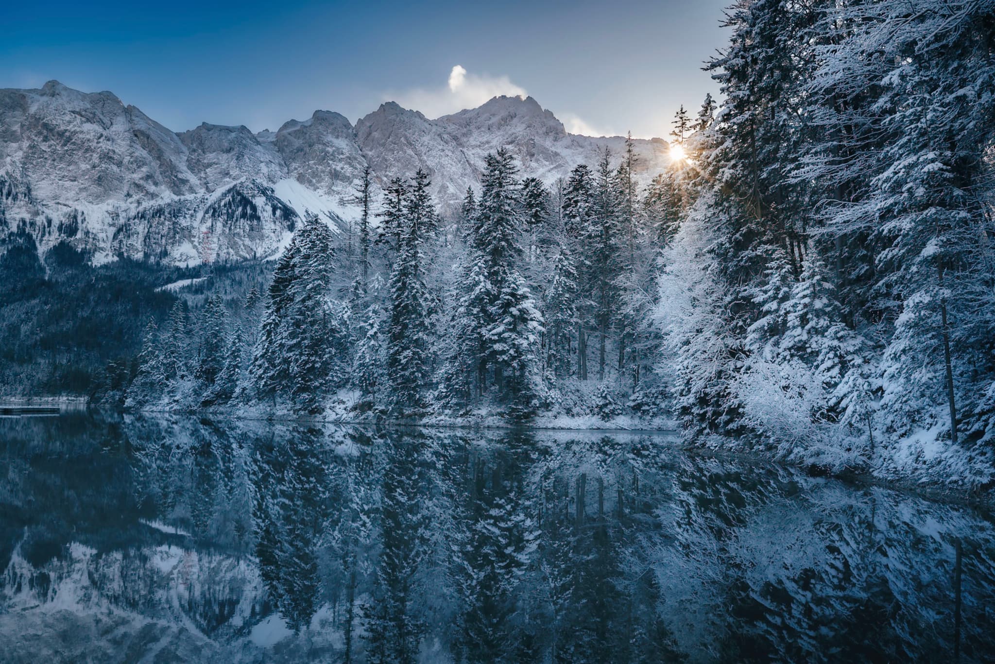 A serene winter landscape with snow-covered trees reflected in a calm lake, set against a backdrop of majestic mountains and a rising sun