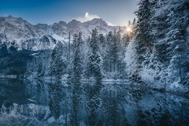 A serene winter landscape with snow-covered trees reflected in a calm lake, set against a backdrop of majestic mountains and a rising sun