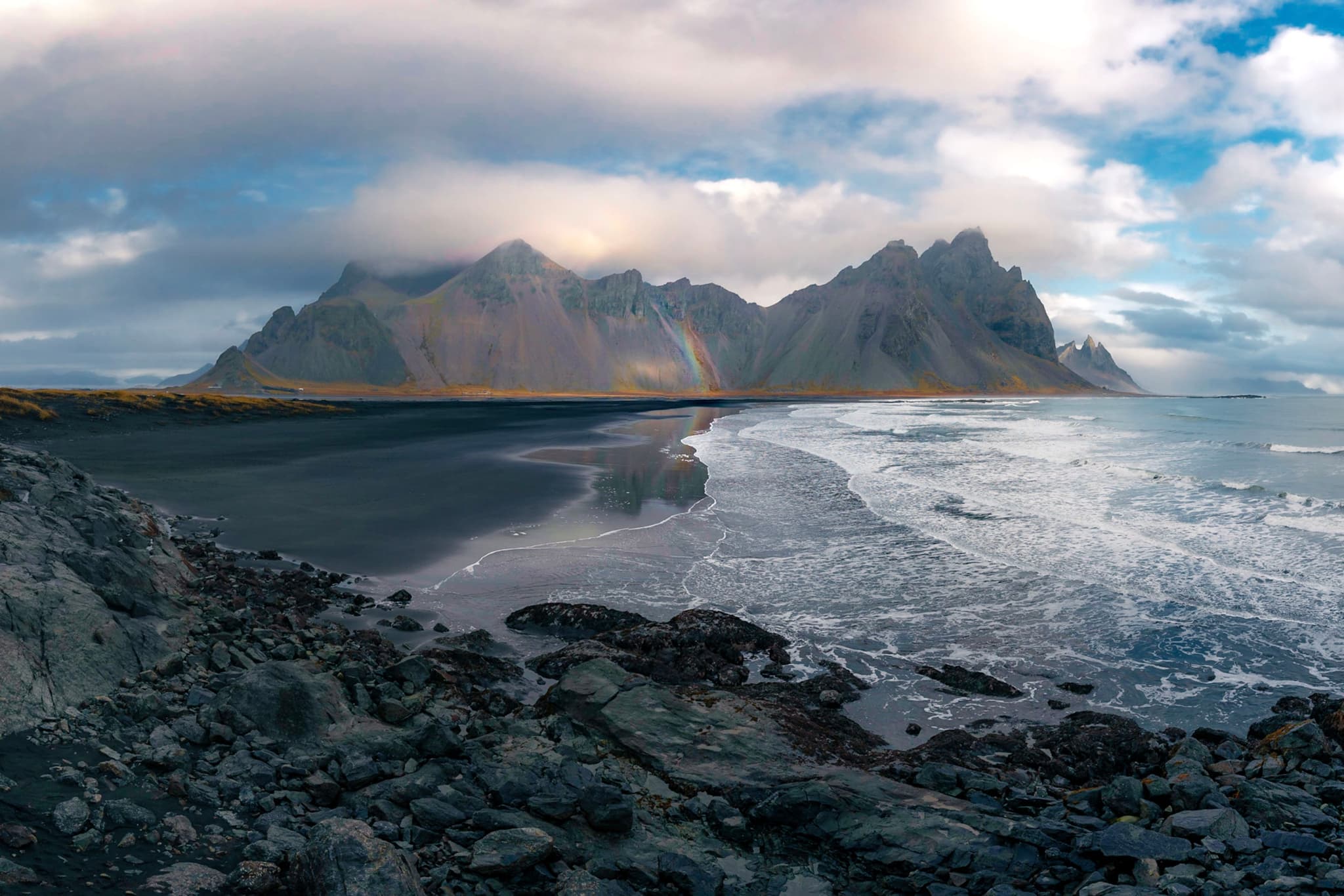 rugged black-sand beach with crashing waves and jagged mountains under a dramatic, cloud-filled sky
