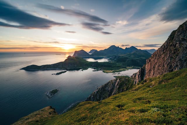 A scenic coastal landscape with mountains, a calm sea, and a sunset casting warm light over the horizon