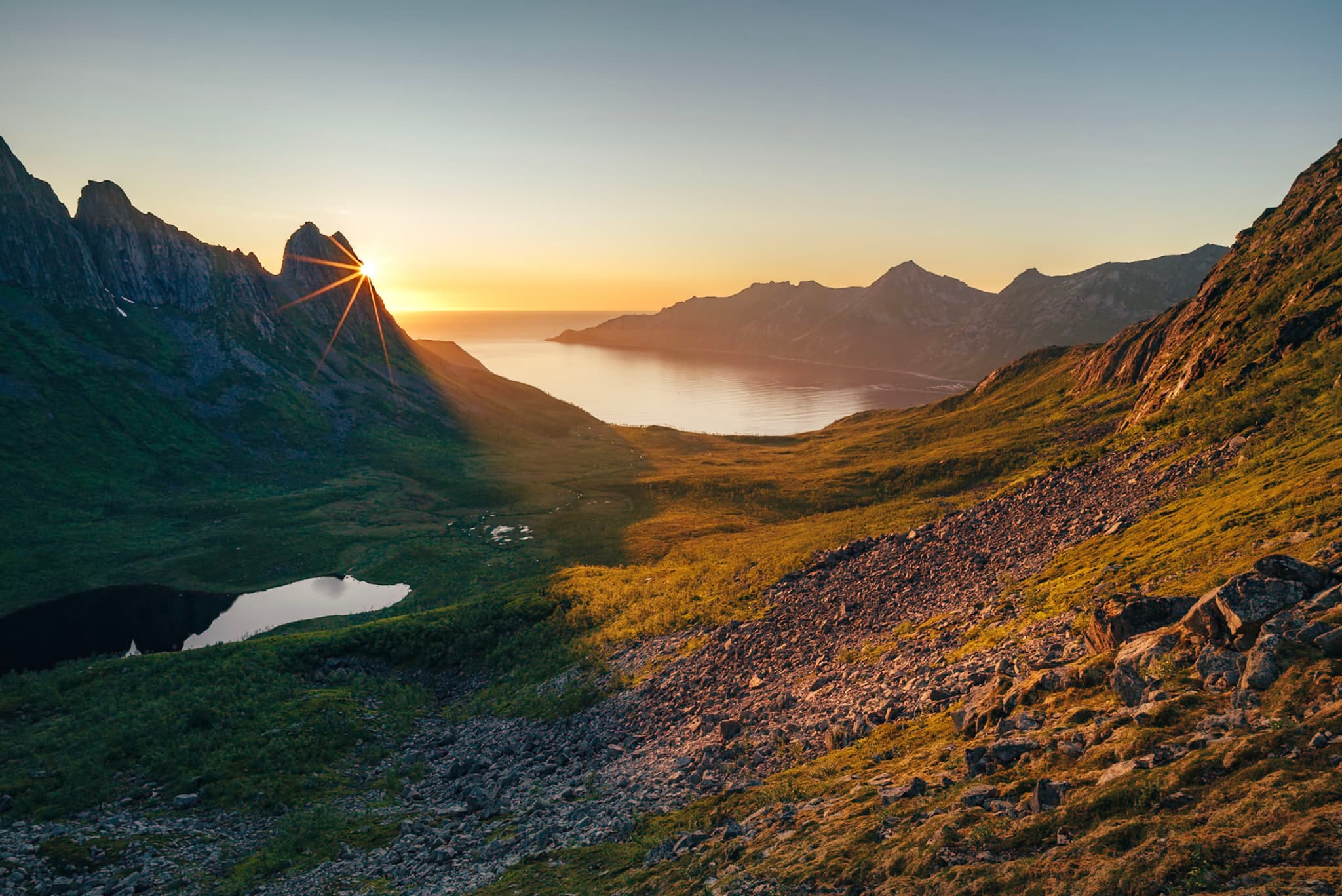 A scenic landscape with a sun setting behind jagged mountains, casting a warm glow over a valley with a small lake and grassy slopes