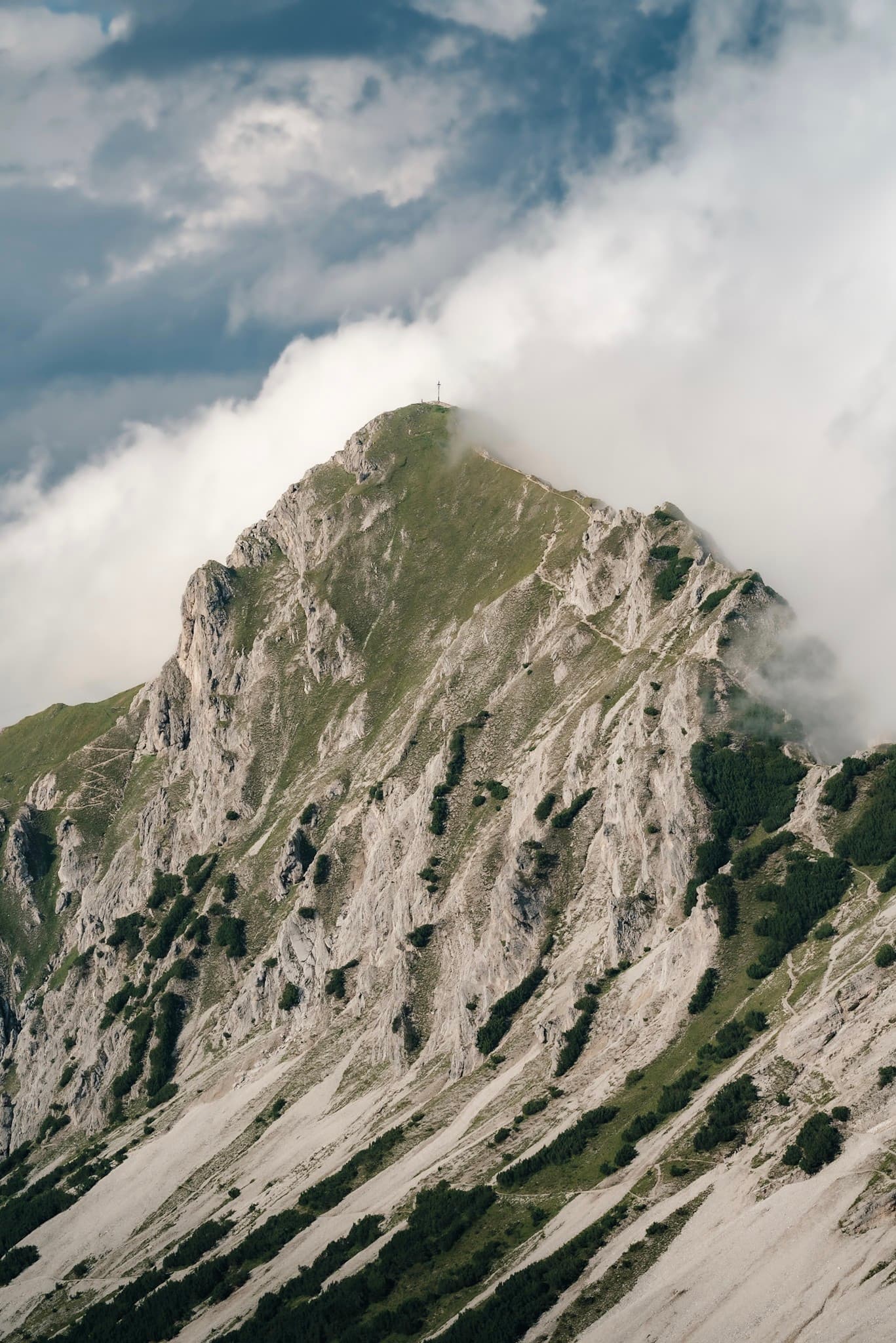 Steep jagged mountain peak with patches of green grass and rock rising into swirling clouds under a partly cloudy sky