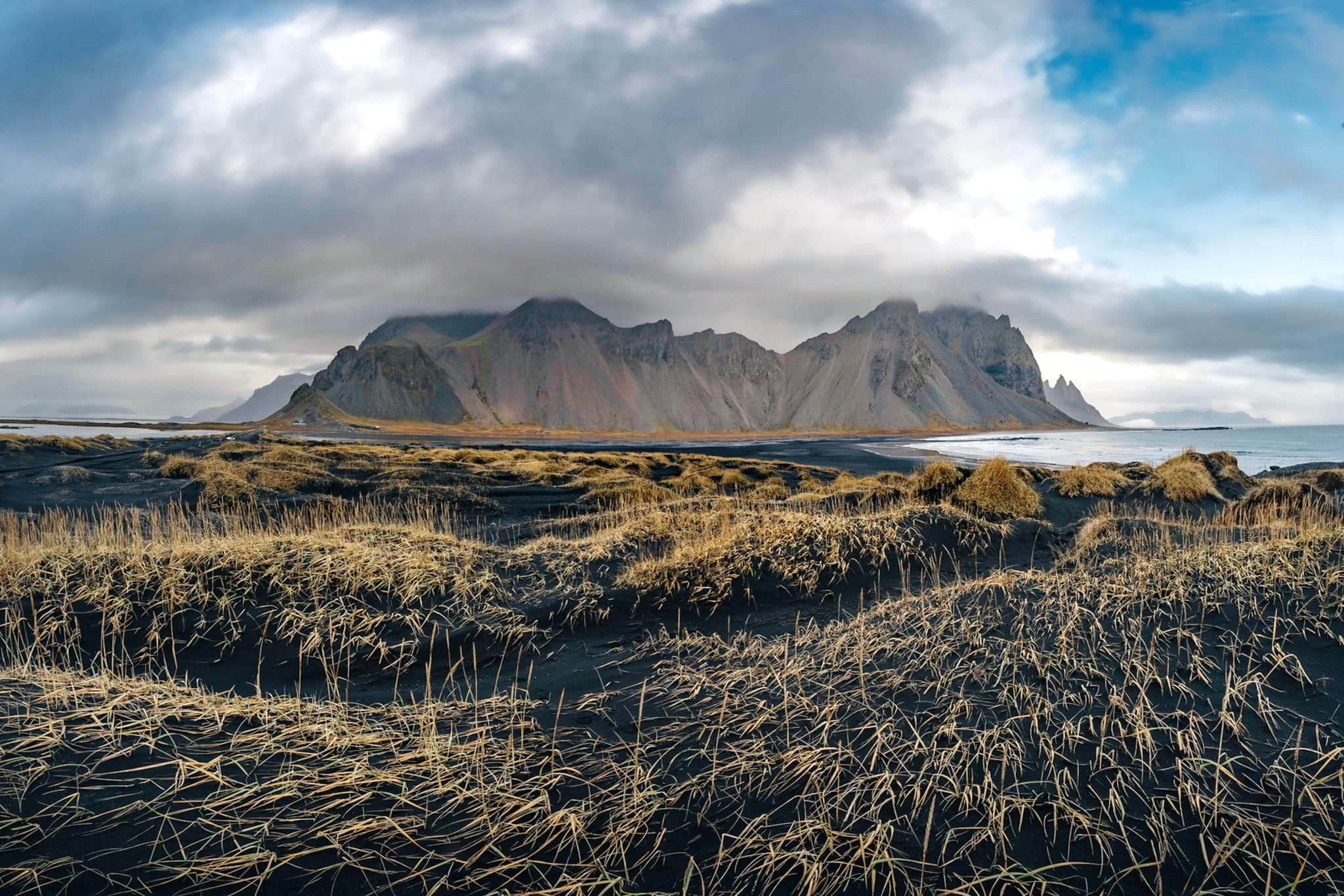 A dramatic landscape featuring rugged mountains under a cloudy sky, with a foreground of dark sand and sparse grass