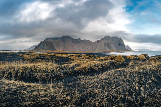 A dramatic landscape featuring rugged mountains under a cloudy sky, with a foreground of dark sand and sparse grass