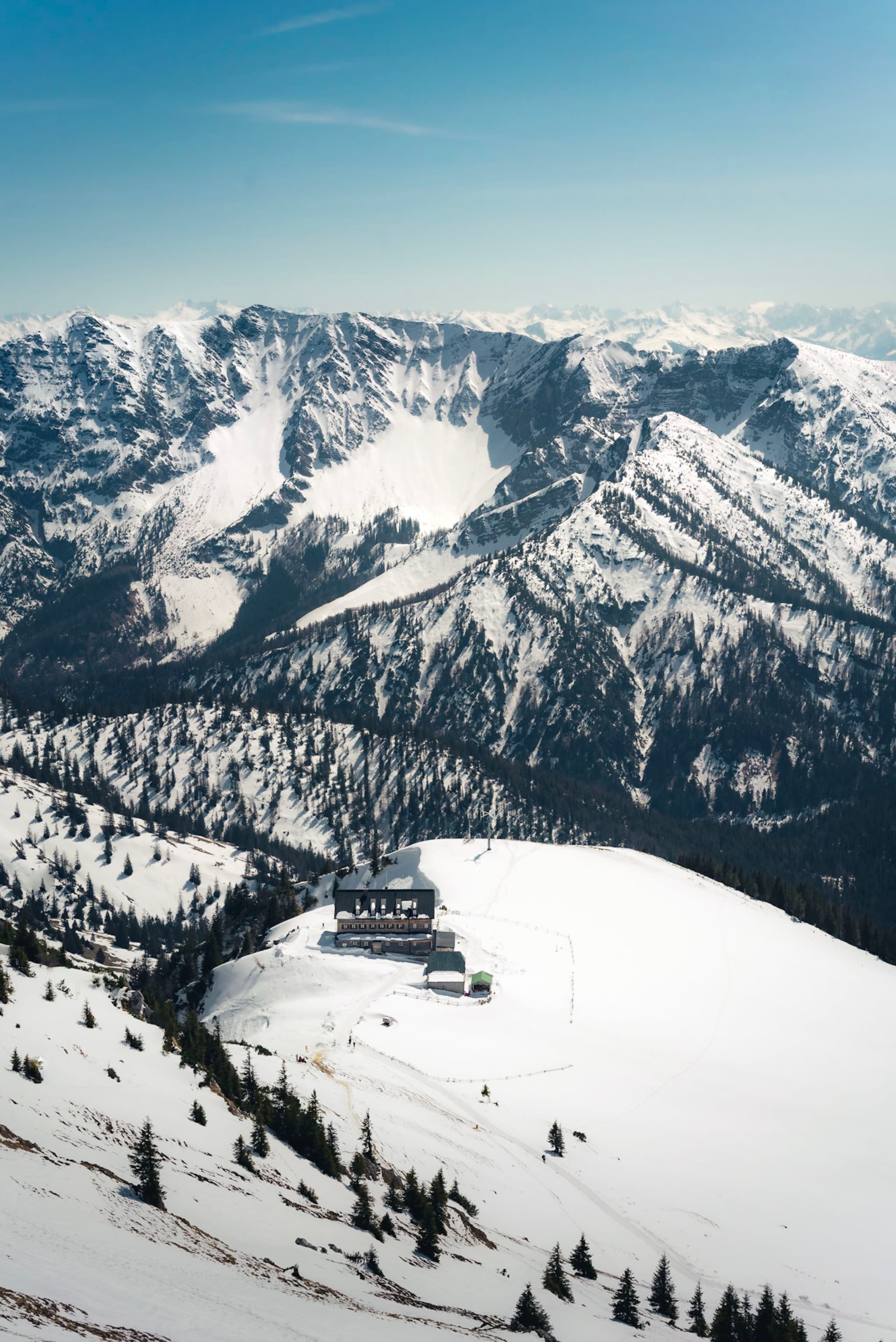 Snow-covered alpine mountains with a small lodge and scattered trees on a high ridge under a clear blue sky