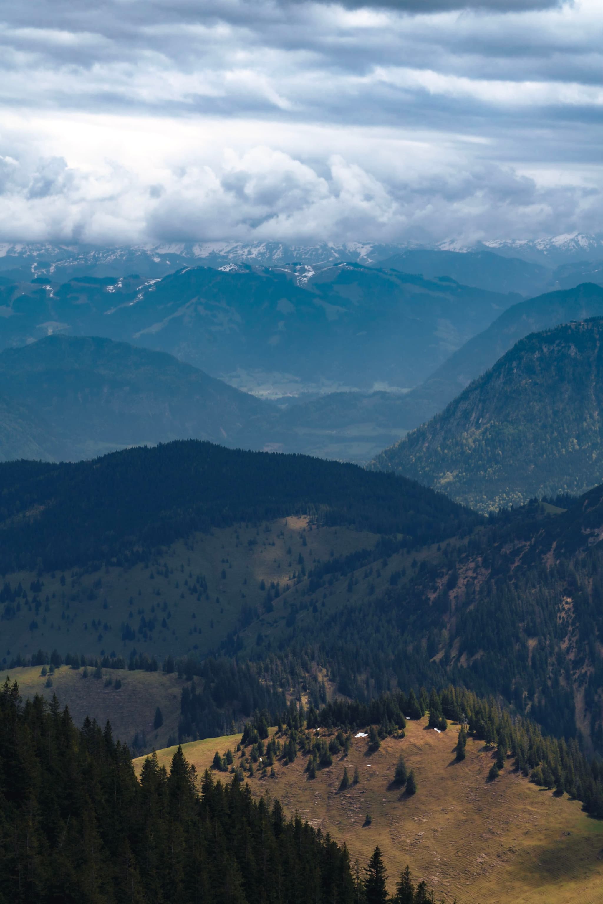 Layered mountain ridges under a cloudy sky with a sunlit grassy slope and scattered trees in the foreground