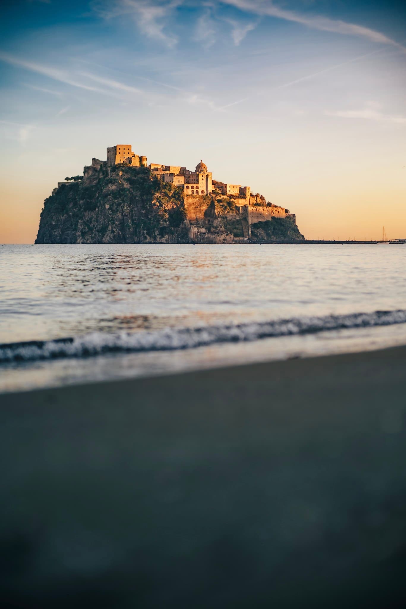 Fortified castle perched on a rocky island at sunset, viewed from a calm shoreline with gentle waves in the foreground