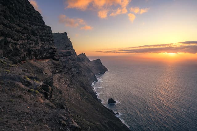 Steep rocky cliffs descending into a calm sea at sunset under a colorful cloud-streaked sky