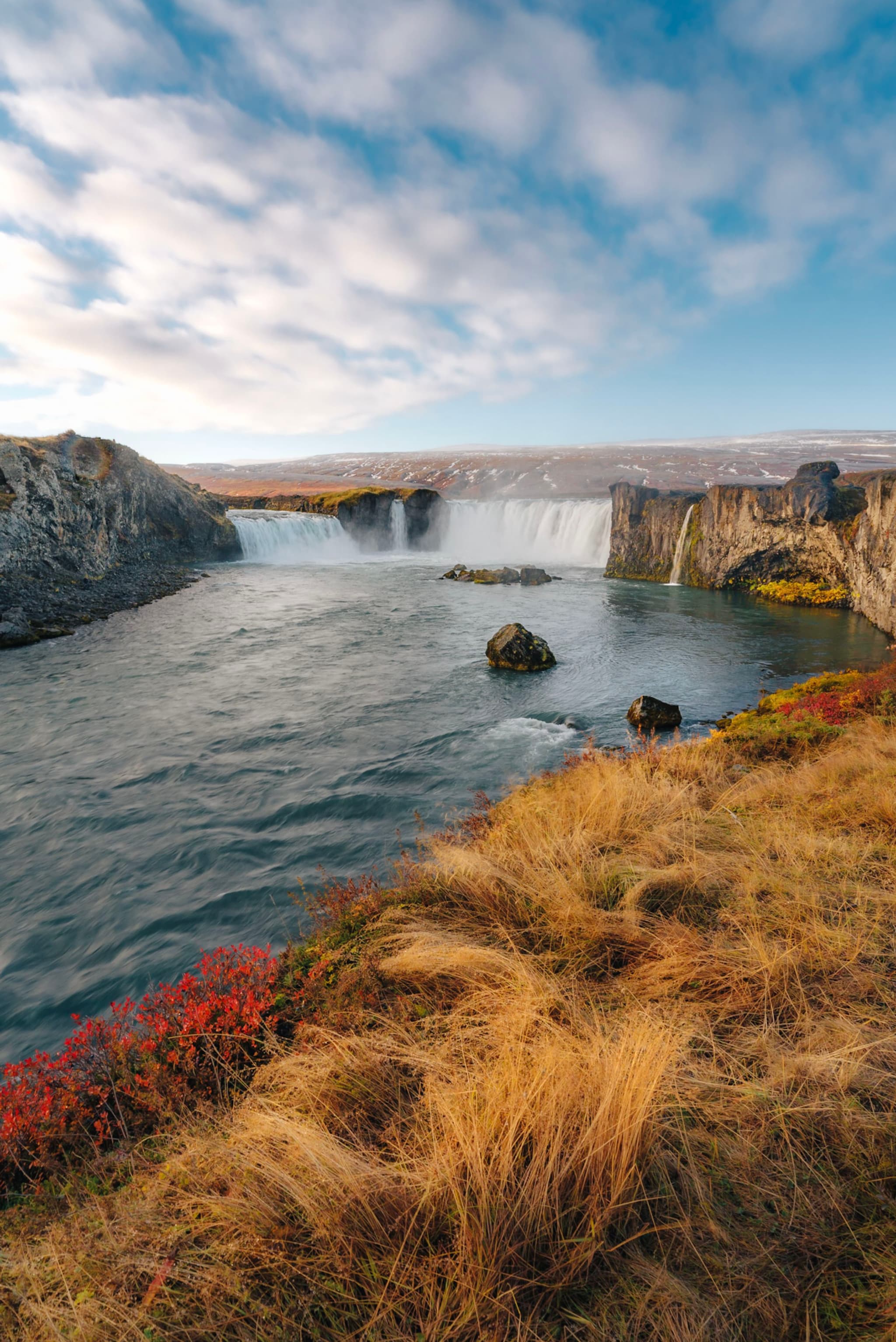 Wide river rushing toward a broad waterfall, bordered by golden grasses and low red shrubs under a partly cloudy sky