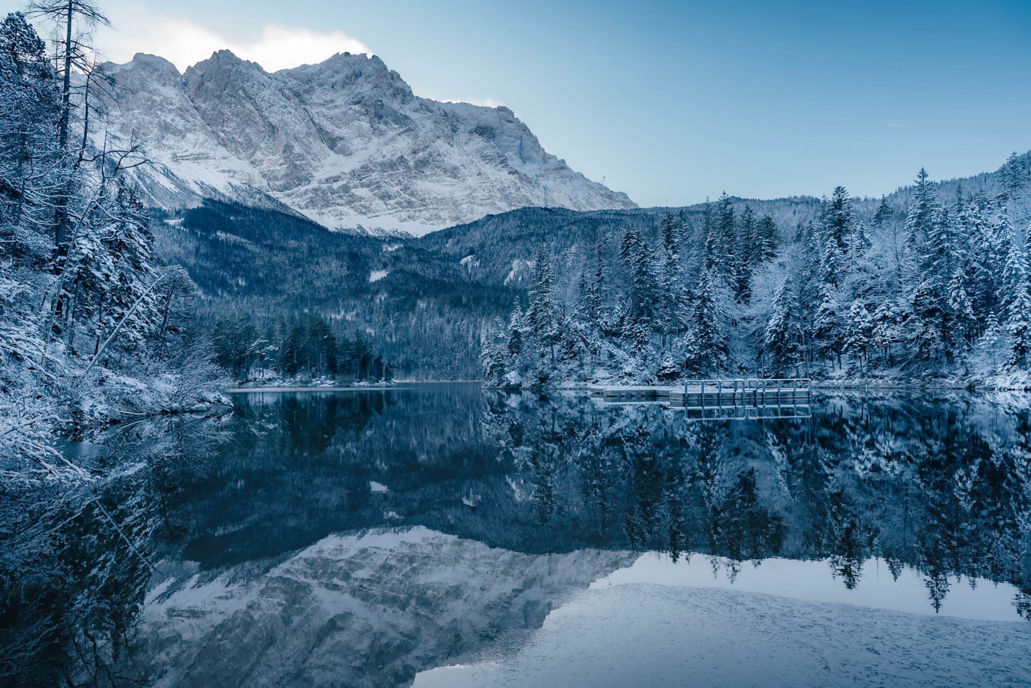 A serene winter landscape with snow-covered mountains and trees reflected in a calm lake