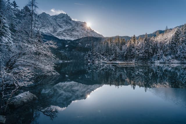 A serene mountain landscape with snow-covered peaks and trees reflected in a calm lake under a clear blue sky