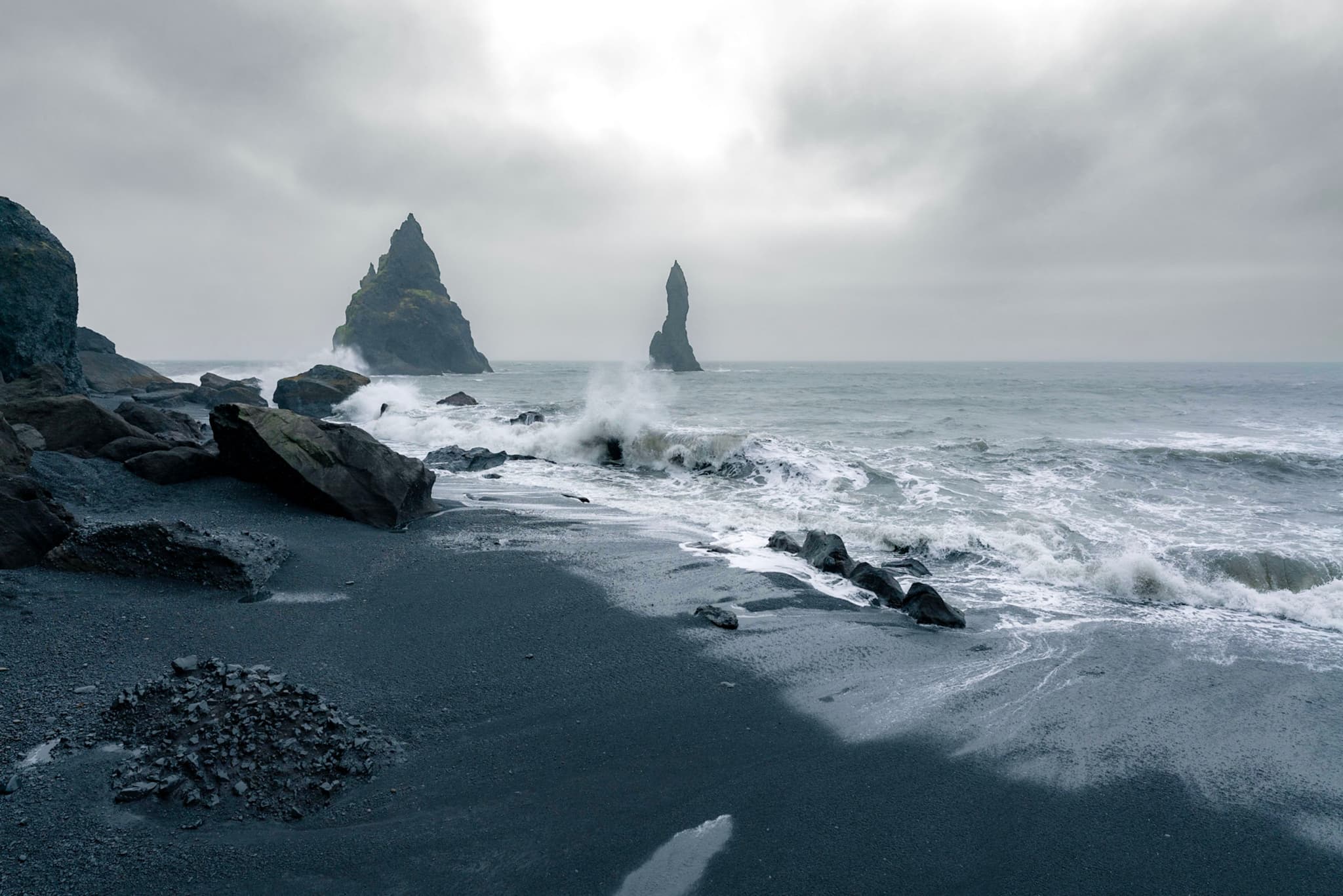 Waves crash onto a dark rocky shoreline with jagged sea stacks rising from the misty ocean under an overcast sky