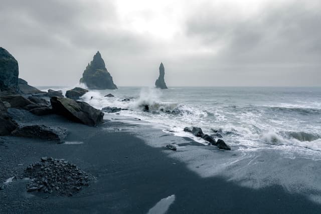 Waves crash onto a dark rocky shoreline with jagged sea stacks rising from the misty ocean under an overcast sky
