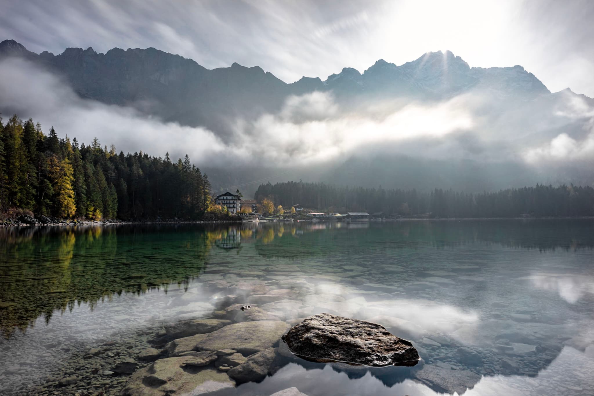 Calm lake with rocks in the foreground reflecting misty forested mountains under a bright hazy sky